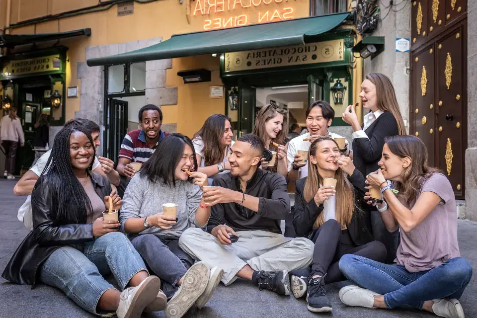 Students sit in front of a store called Chocolateria San Gines dunking pastries in cups of hot chocolate and eating them.
